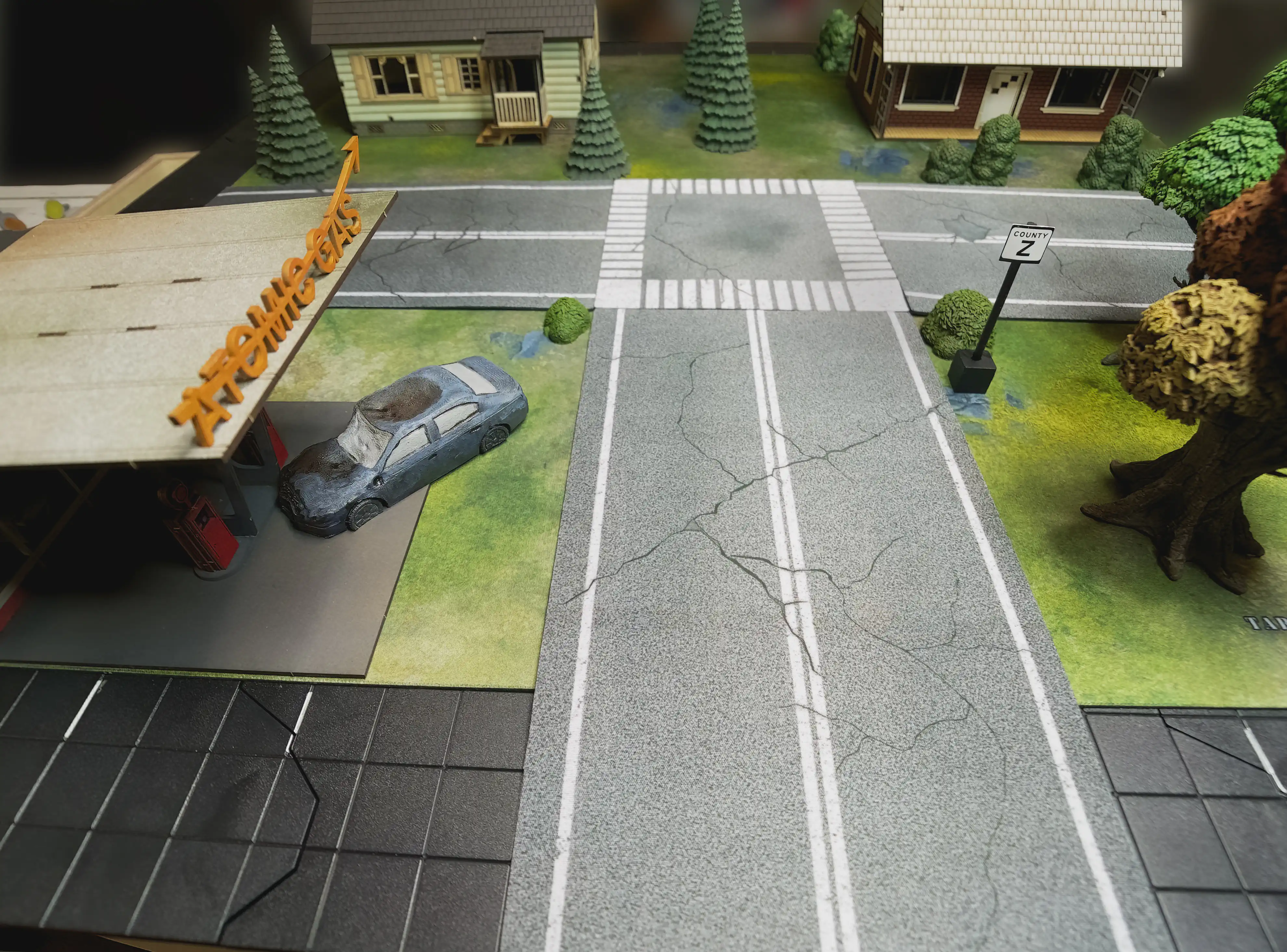 A photo of the game board taken just above eye level for the miniatures. The photo looks down the street toward the top of a T-junction. There is a gas station on the left, woods on the right, and two houses at the end of the road.
